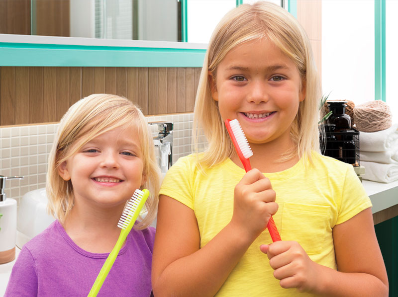 Two young girls in a bathroom, smiling and holding giant toothbrushes.